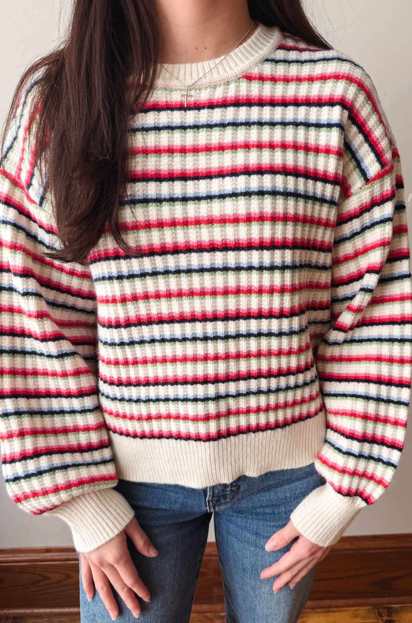A person wearing a chunky knit sweater with horizontal stripes in cream, red, and navy blue, paired with blue jeans.