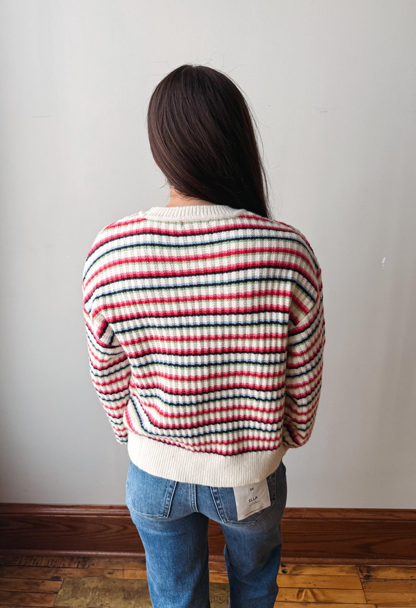 A person viewed from behind wearing a striped knit sweater with red, black, and white horizontal stripes, paired with blue jeans, standing against a plain light gray wall.