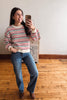 A woman taking a mirror selfie in a room with a light-colored wall and wooden floor, wearing a red, white, and black striped long-sleeve knit sweater, blue jeans, and brown boots.
