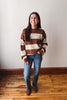 A woman wearing a brown and cream striped knit sweater, blue jeans, and brown boots, standing against a plain off-white wall with a wooden floor.