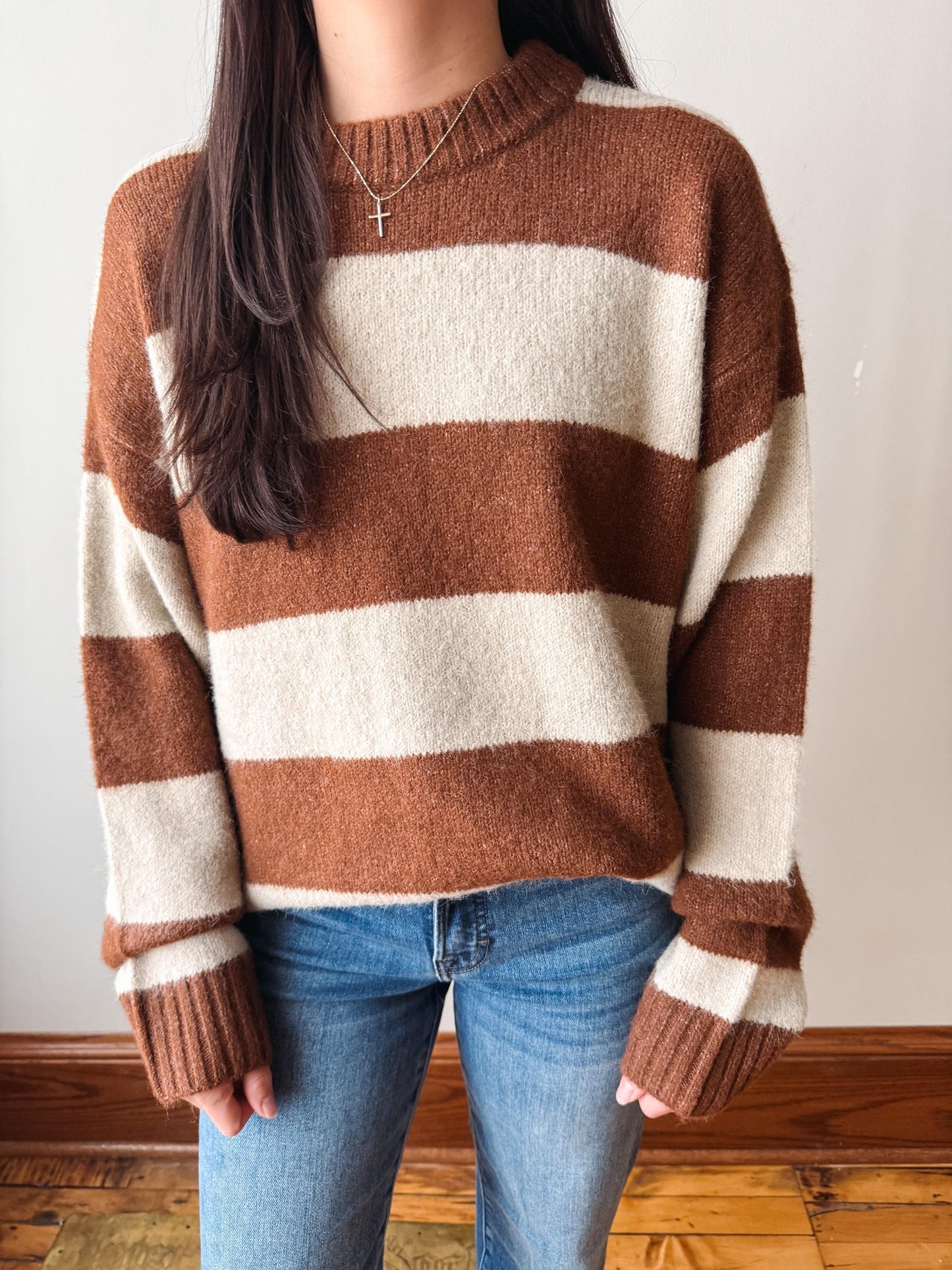 A person wearing a fuzzy knit sweater with wide brown and cream horizontal stripes, paired with blue jeans, standing indoors against a plain light wall.