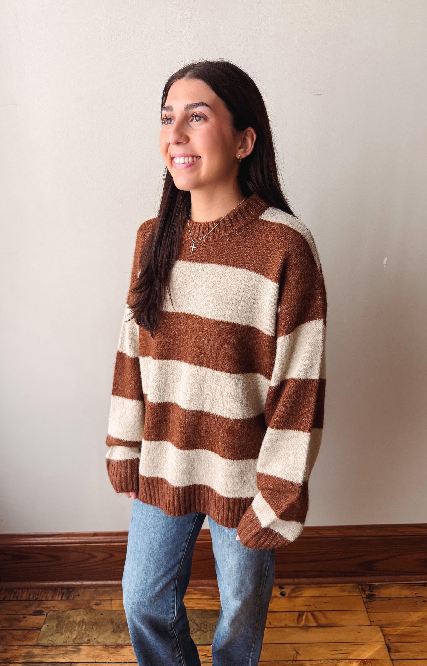 A woman wearing a brown and cream striped knit sweater and blue jeans, standing against a plain off-white wall with a wooden baseboard and floor.