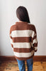 A person viewed from behind wearing a brown and cream striped knit sweater and blue jeans, standing against a plain light wall on a wooden floor.