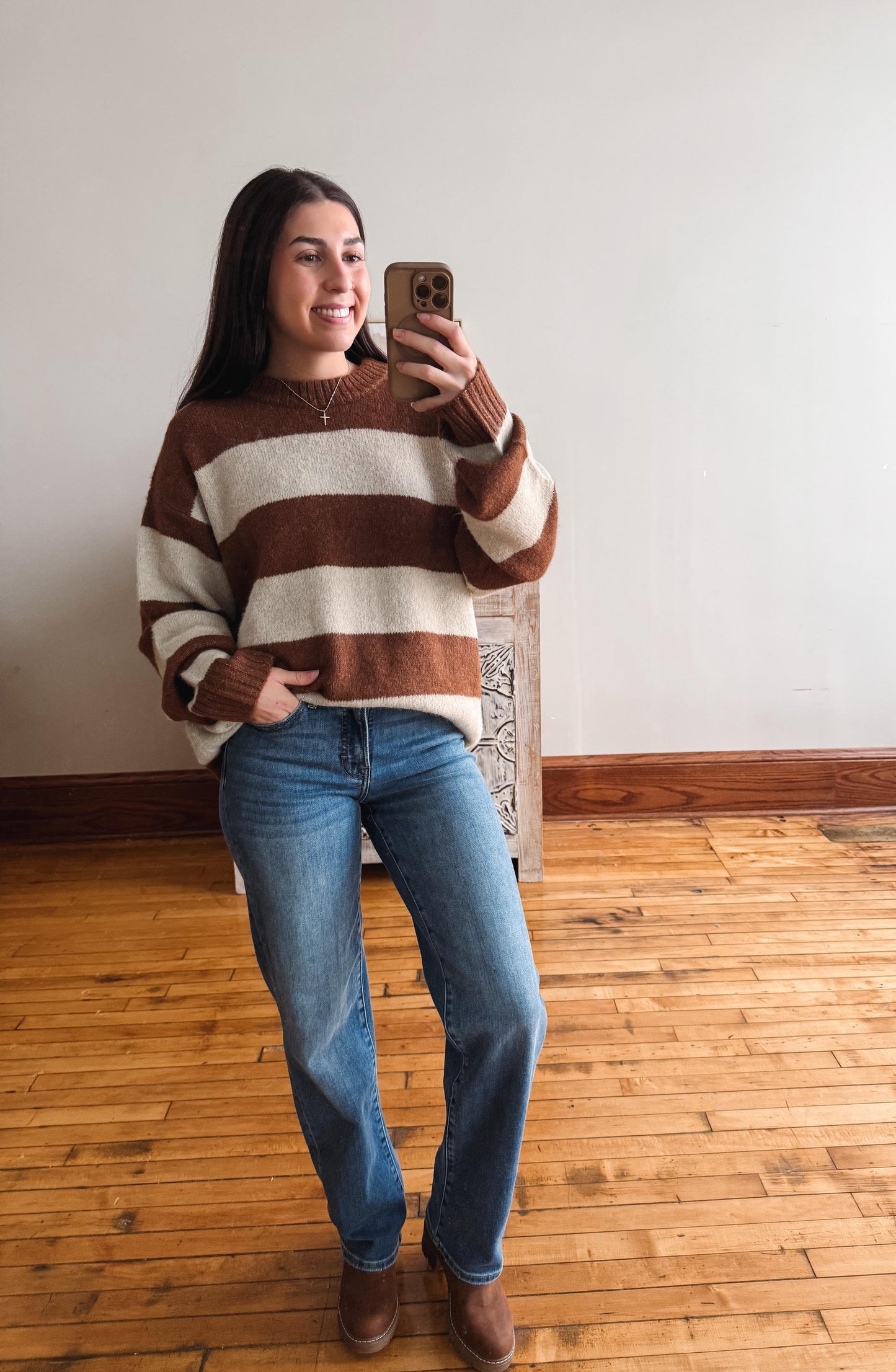 A woman takes a mirror selfie wearing a brown and cream striped knit sweater, blue jeans, and brown boots in a room with a light wall and wooden floor.