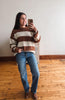 A woman takes a mirror selfie wearing a brown and cream striped knit sweater, blue jeans, and brown boots in a room with a light wall and wooden floor.
