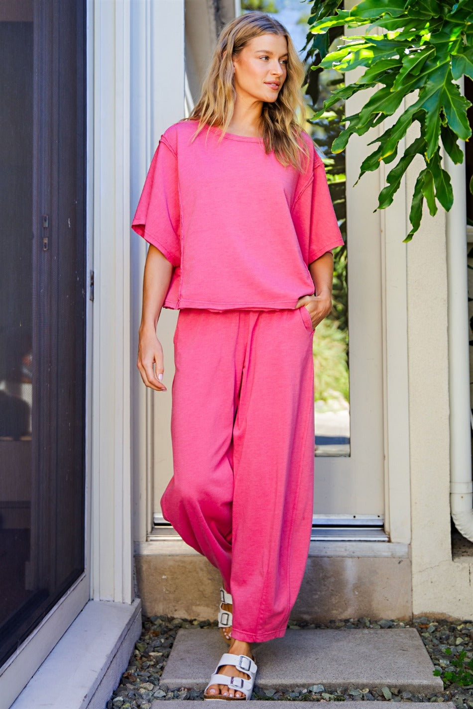 Woman wearing a bright pink outfit standing in front of a glass door with greenery around.