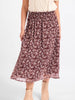 A woman wearing a mid-length, flowy skirt with a dense small floral print in dark brown and white. The skirt has an elastic waistband and is styled with a cream-colored knit top and beige ankle boots against a plain white studio background.