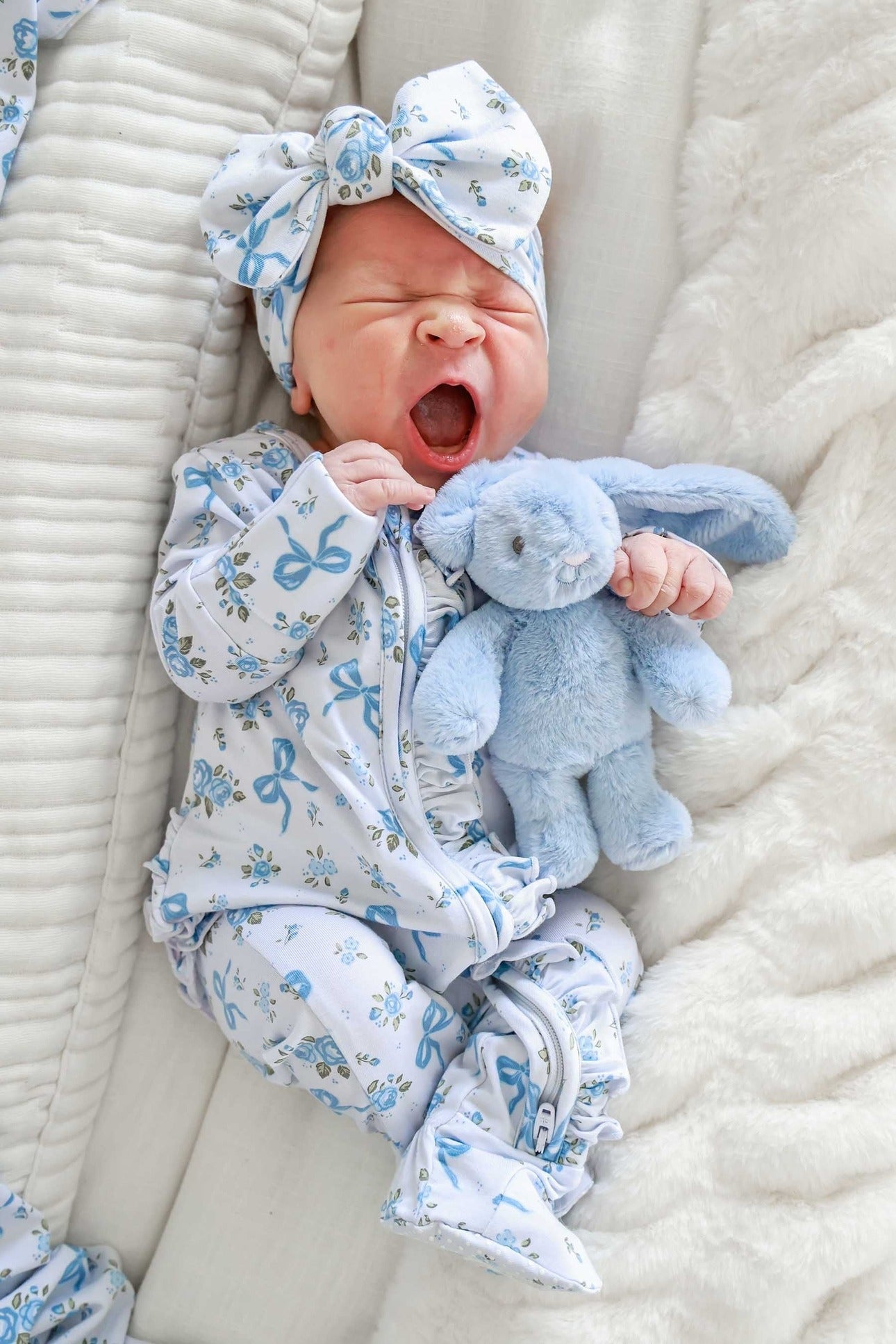 A newborn baby wearing a light blue and white patterned footed sleeper and matching bow headband, holding a light blue plush bunny toy while lying on a soft cream blanket.