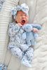 A newborn baby wearing a light blue and white patterned footed sleeper and matching bow headband, holding a light blue plush bunny toy while lying on a soft cream blanket.