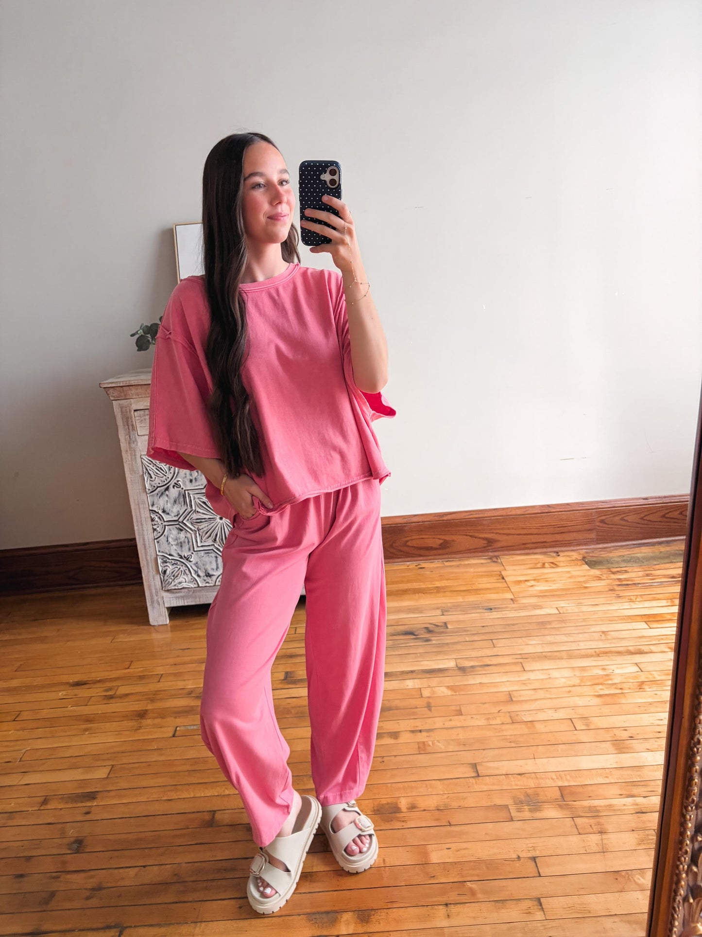Person wearing a pink outfit taking a mirror selfie in a room with wooden flooring.
