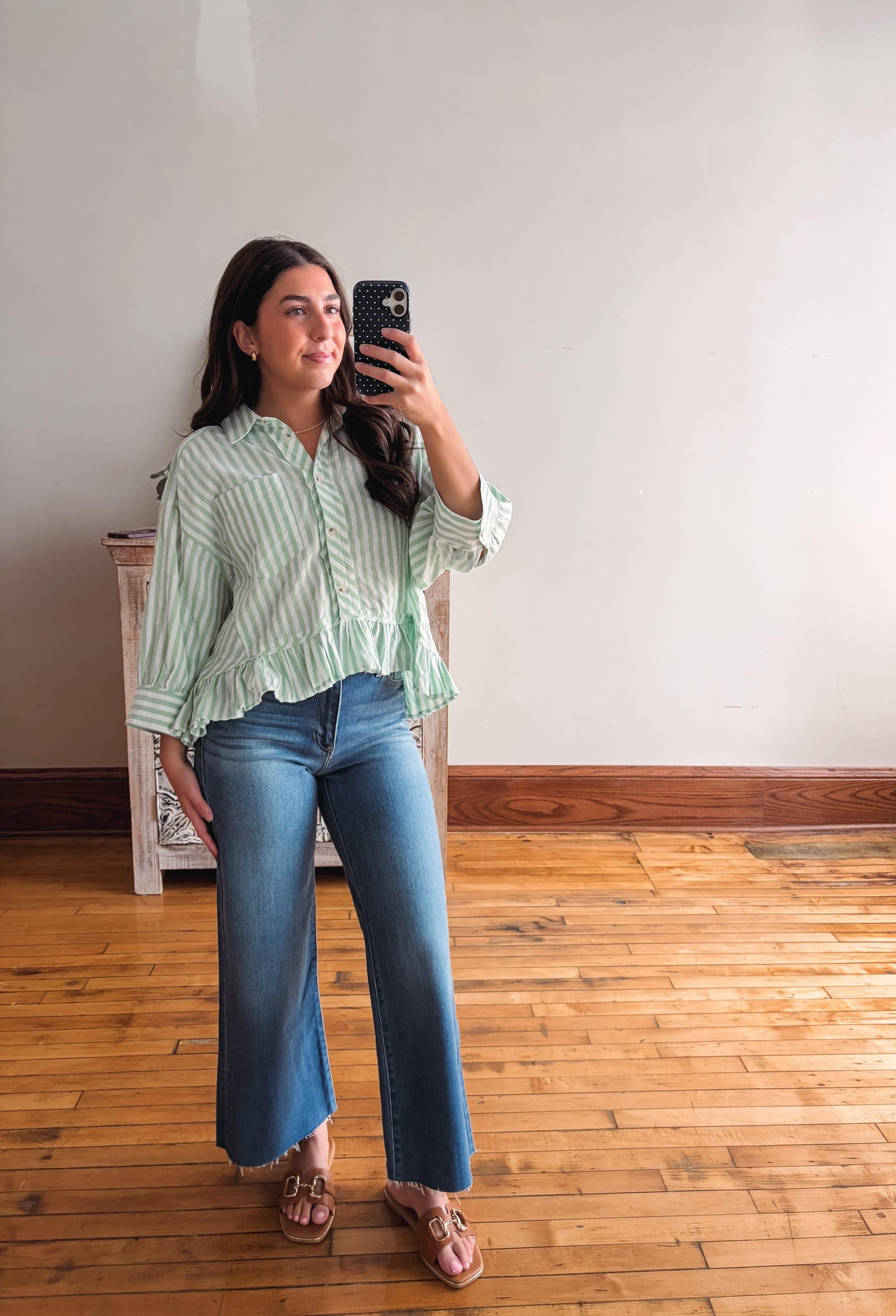 Woman taking a mirror selfie wearing a green striped shirt and blue jeans on a wooden floor.