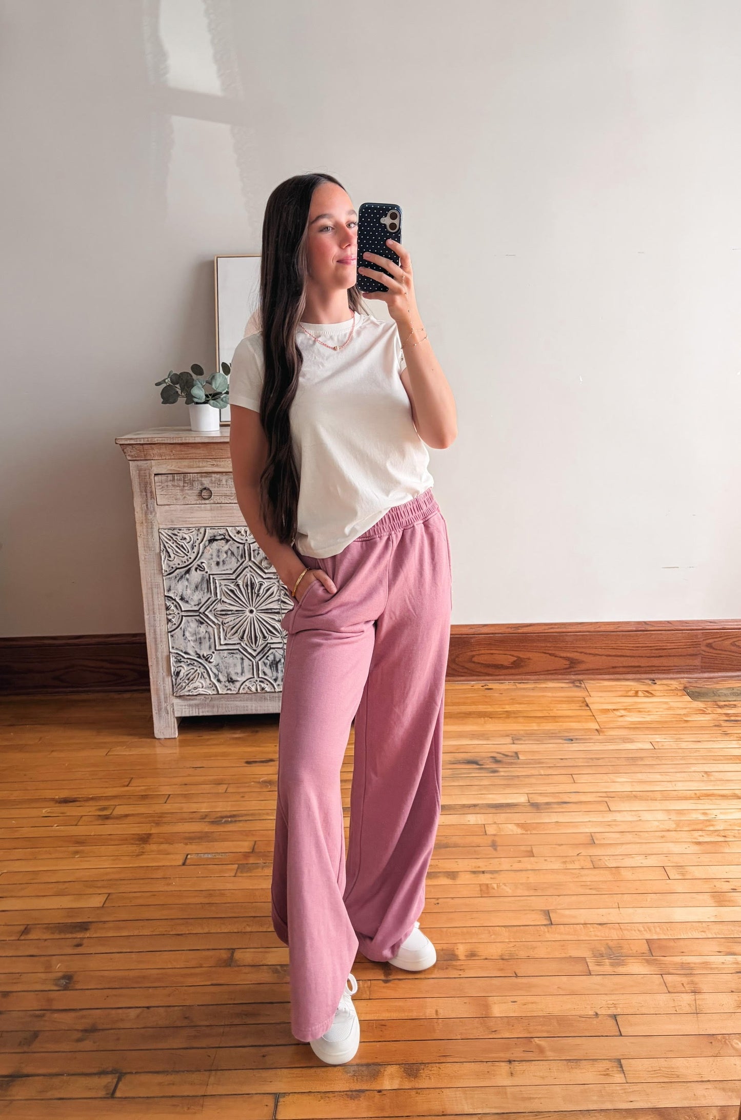 Woman taking a mirror selfie wearing a white top and pink pants in a room with wooden flooring and a decorative cabinet.