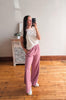 Woman taking a mirror selfie wearing a white top and pink pants in a room with wooden flooring and a decorative cabinet.