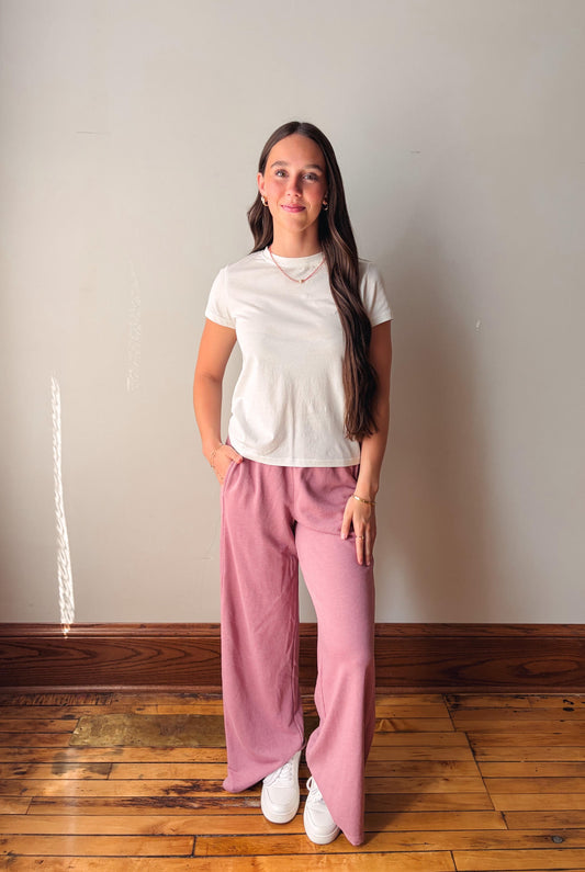 Woman wearing a white t-shirt and pink pants standing against a plain wall.