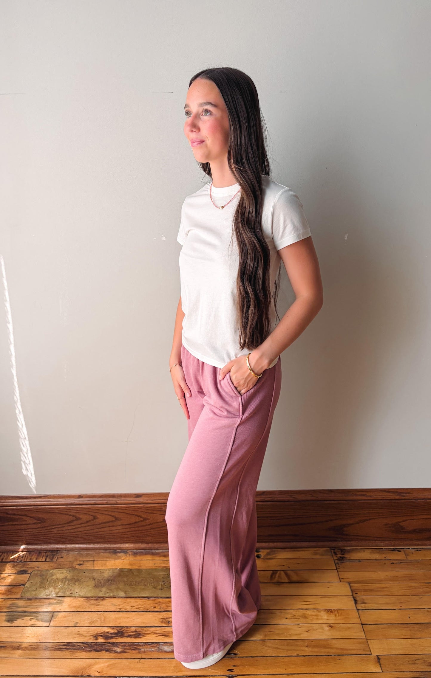 Woman wearing a white t-shirt and pink pants standing against a plain wall.