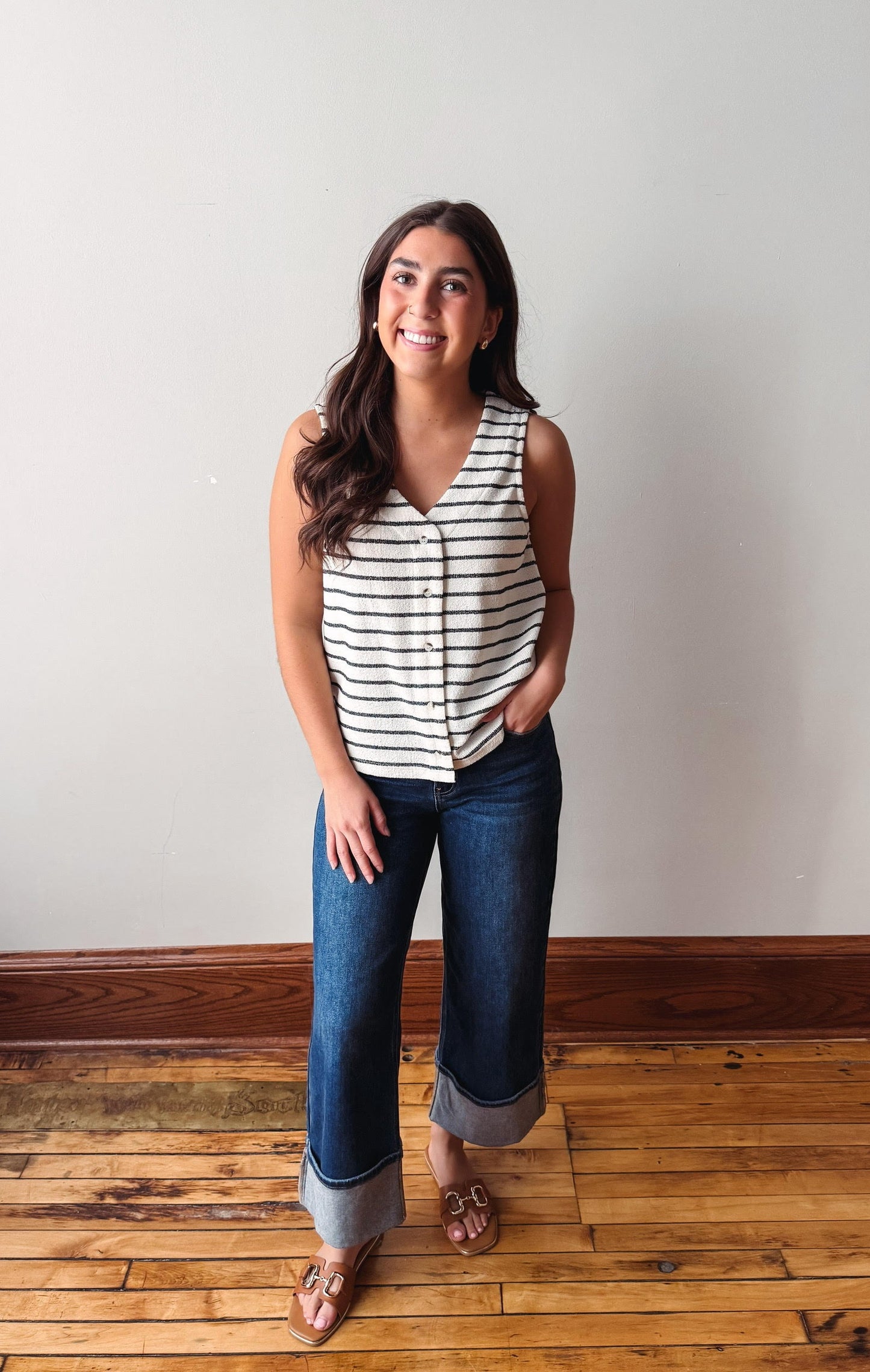 Woman wearing a striped tank top and jeans standing on a wooden floor with a white wall background