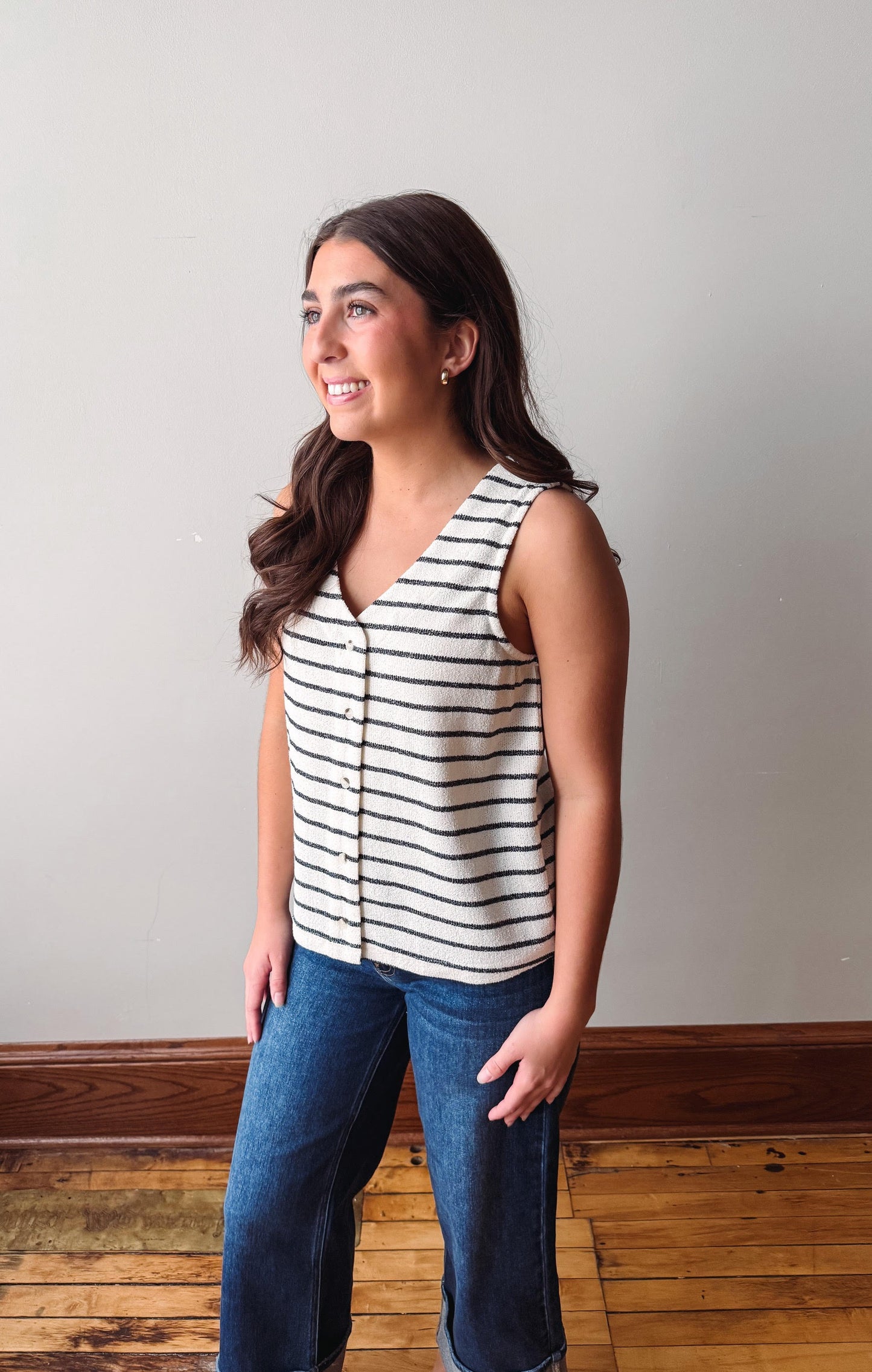 Woman wearing a striped sleeveless top and jeans standing against a plain wall.