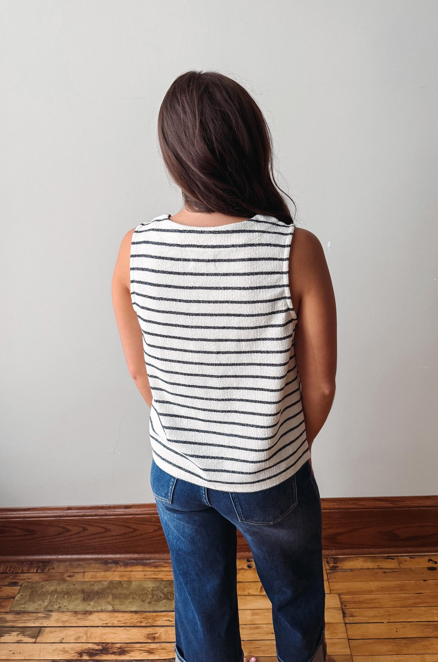 Person wearing a striped sleeveless top and jeans standing against a plain wall.