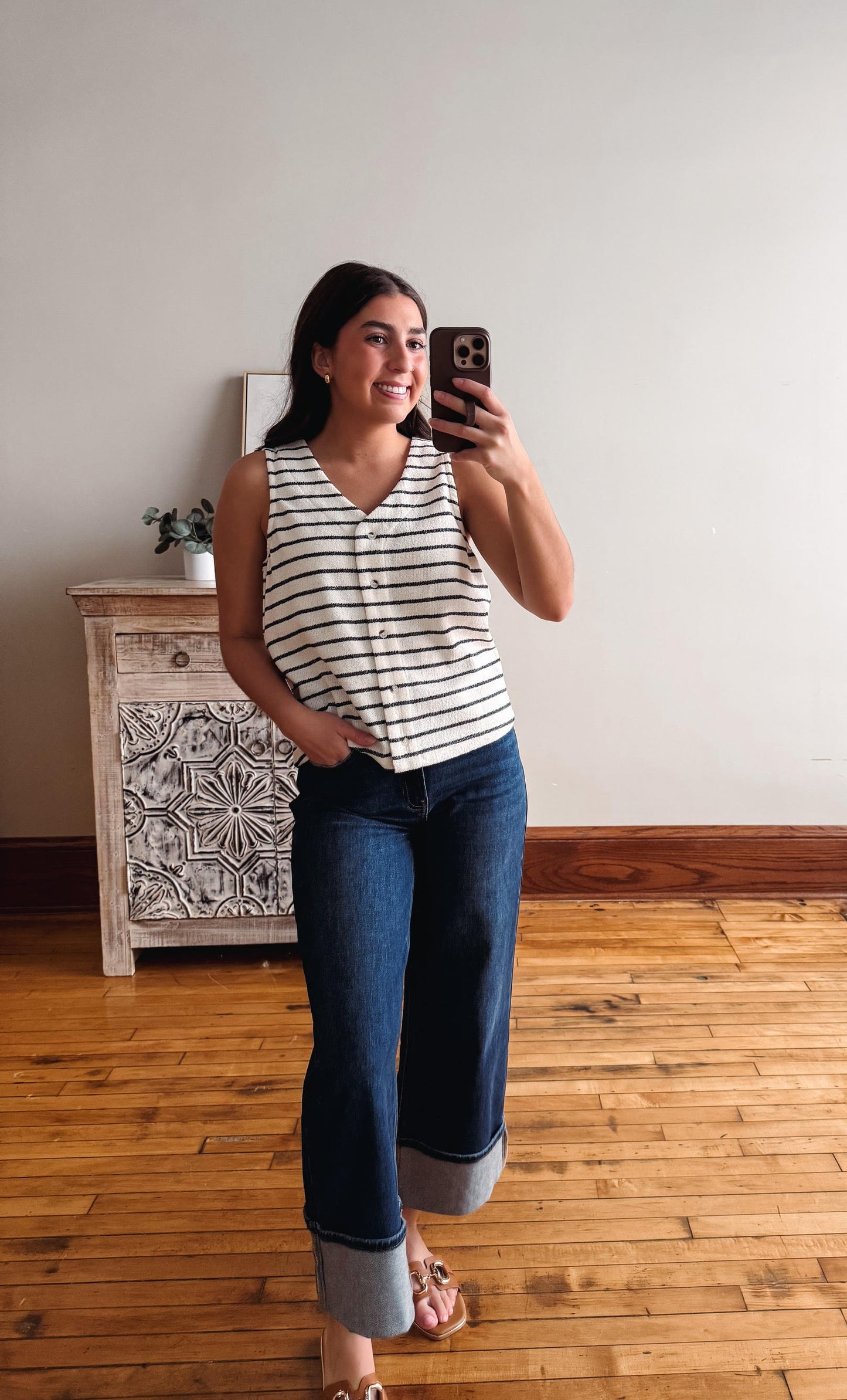 Woman taking a mirror selfie wearing a striped shirt and jeans in a room with wooden flooring and a decorative cabinet.