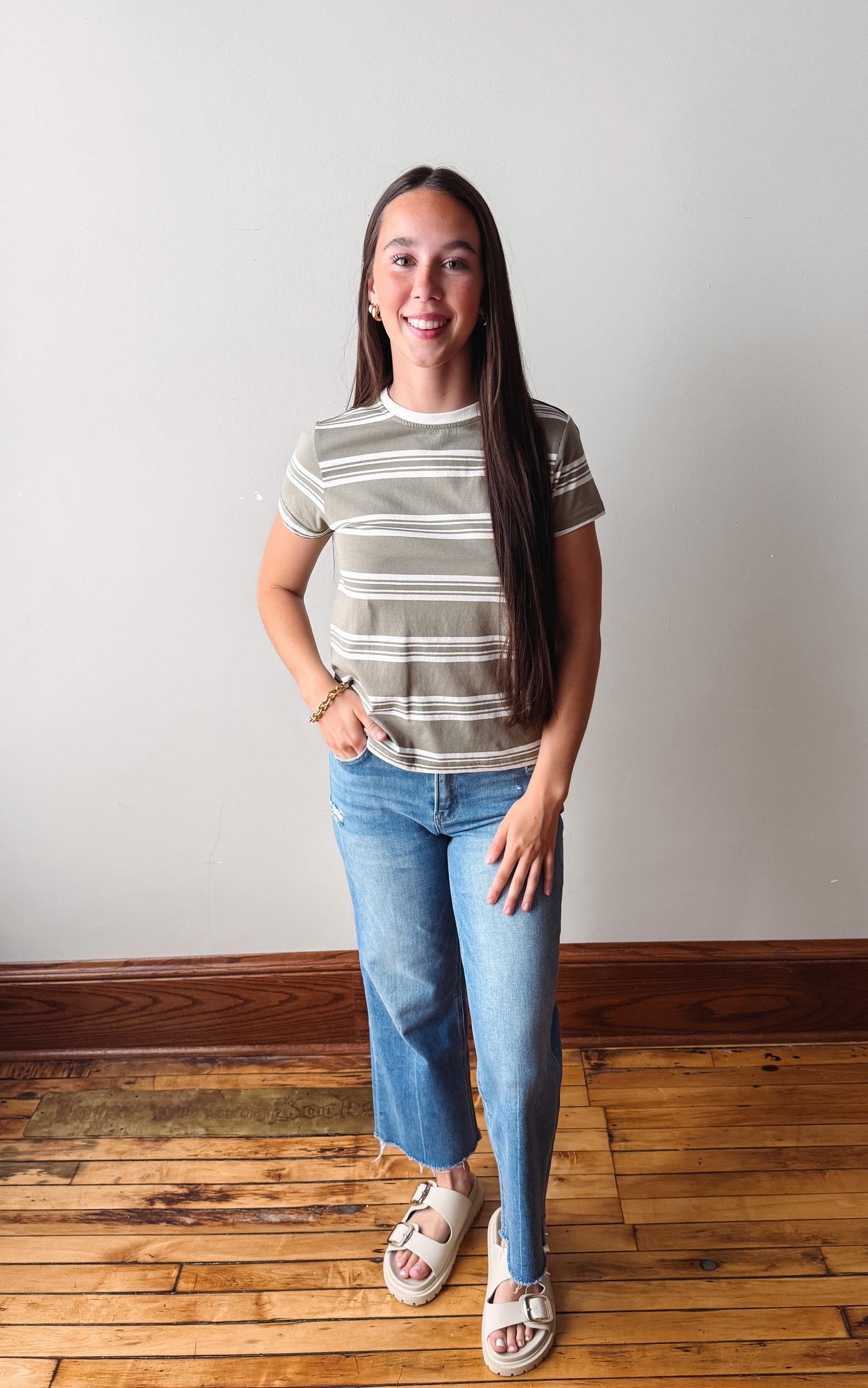 Woman wearing a striped shirt and jeans standing on a wooden floor with a white wall background