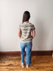Person wearing a striped shirt and blue jeans standing against a plain wall.