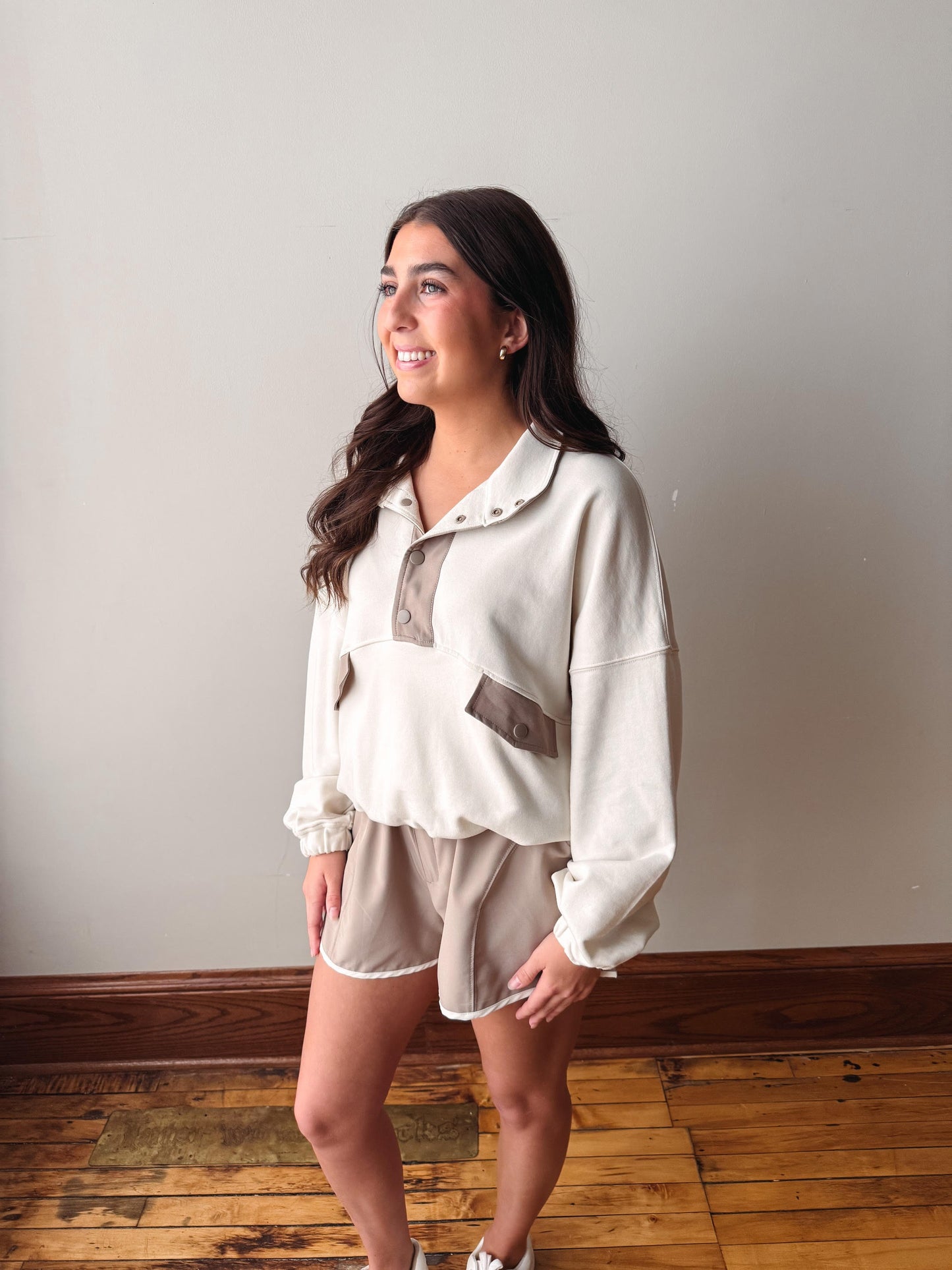 Woman wearing a white blouse and beige shorts standing against a plain wall.
