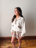 Woman wearing a white blouse and beige shorts standing against a plain wall.