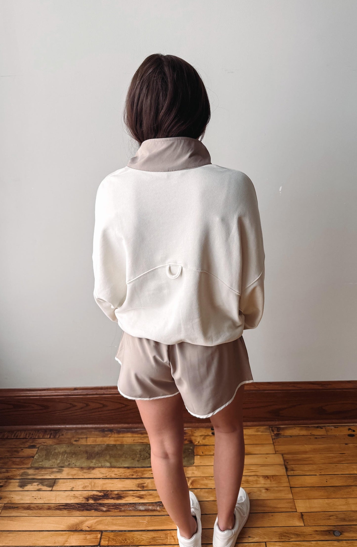 Person wearing a white jacket and beige shorts standing on a wooden floor against a plain wall.