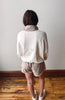 Person wearing a white jacket and beige shorts standing on a wooden floor against a plain wall.