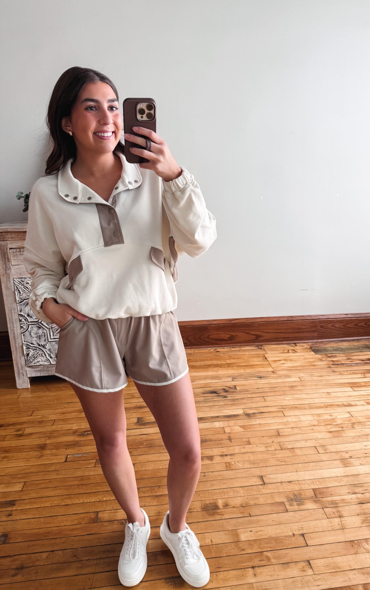 Woman taking a mirror selfie wearing a white blouse and beige shorts on a wooden floor.