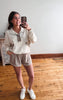 Woman taking a mirror selfie wearing a white blouse and beige shorts on a wooden floor.