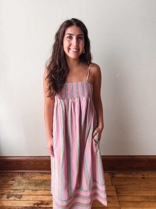 A woman wearing a pink and green striped sleeveless midi dress with a smocked bodice, standing against a plain off-white wall with a wooden floor.