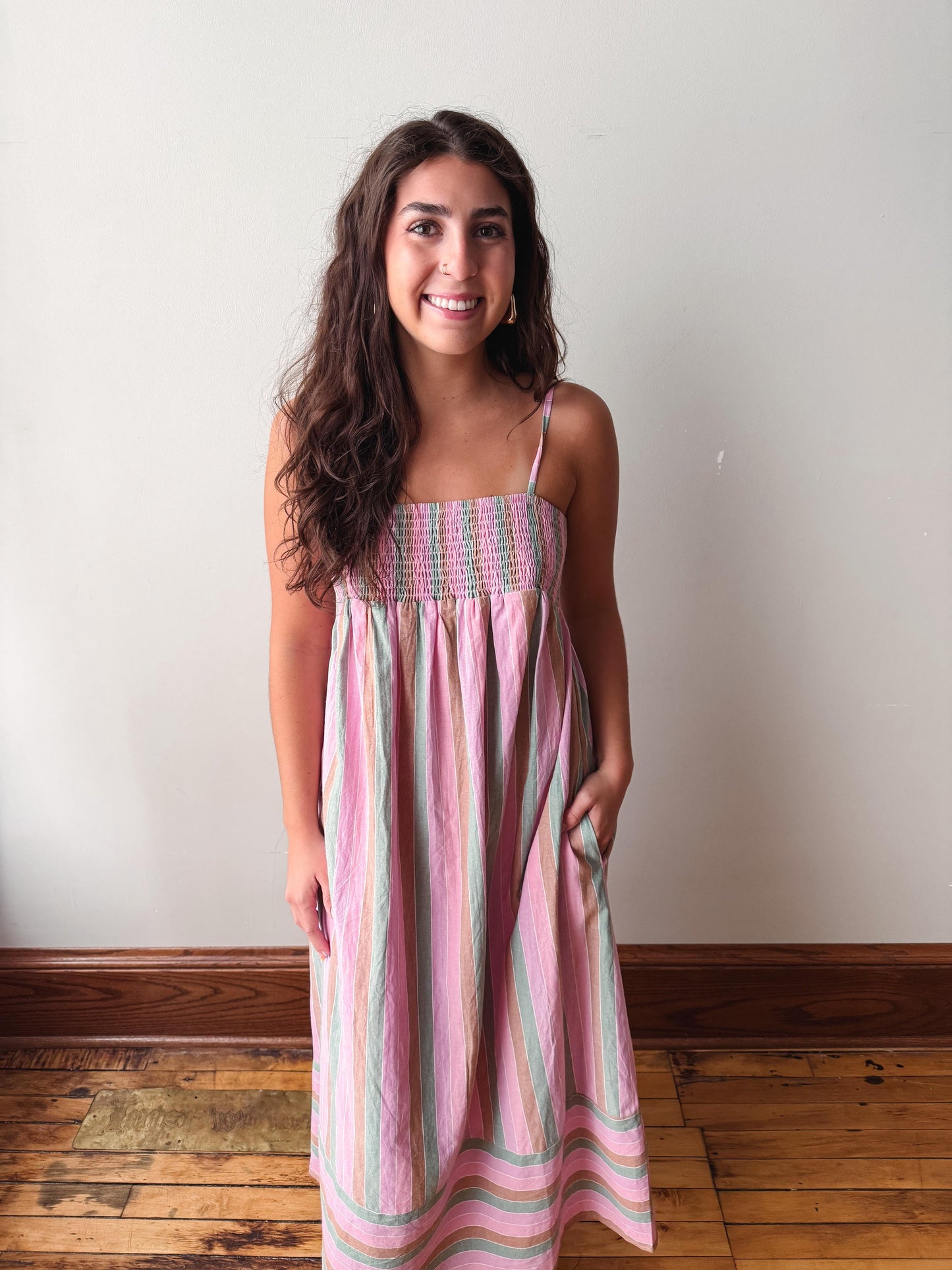 A woman wearing a pink and green striped sleeveless midi dress with a smocked bodice, standing against a plain off-white wall with a wooden floor.