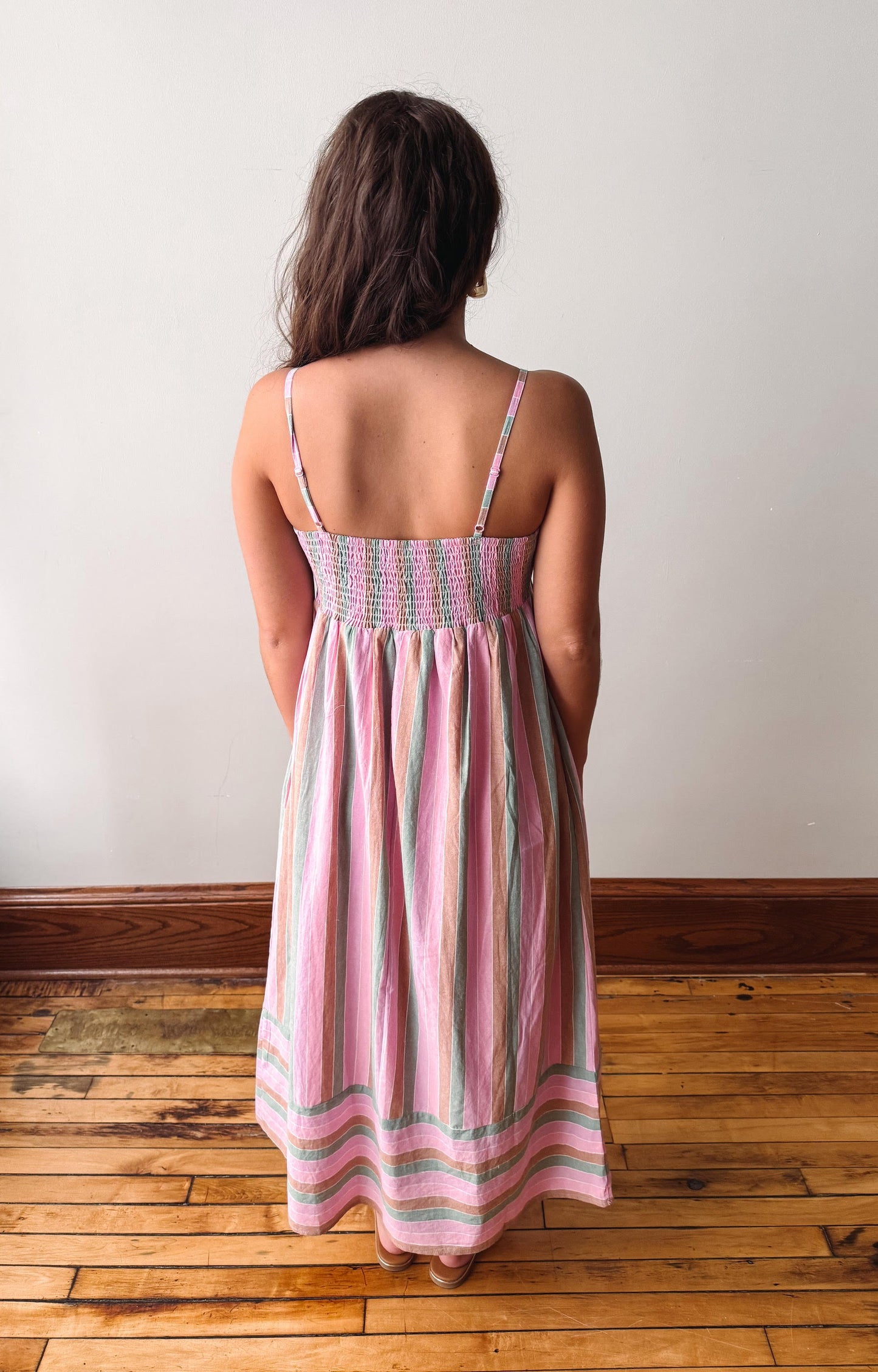 A woman viewed from behind wearing a sleeveless smocked dress with vertical pink, green, and white stripes, standing on a wooden floor against a plain white wall.