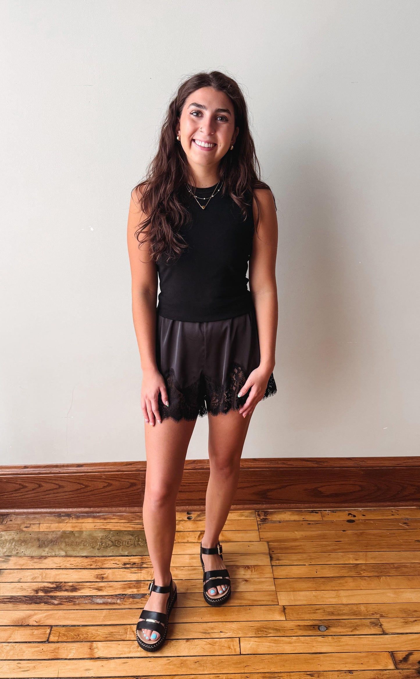 A young woman with long brown hair stands in a room with a light gray wall and wooden floor, wearing a black sleeveless top, black lace-trimmed shorts, and black sandals.