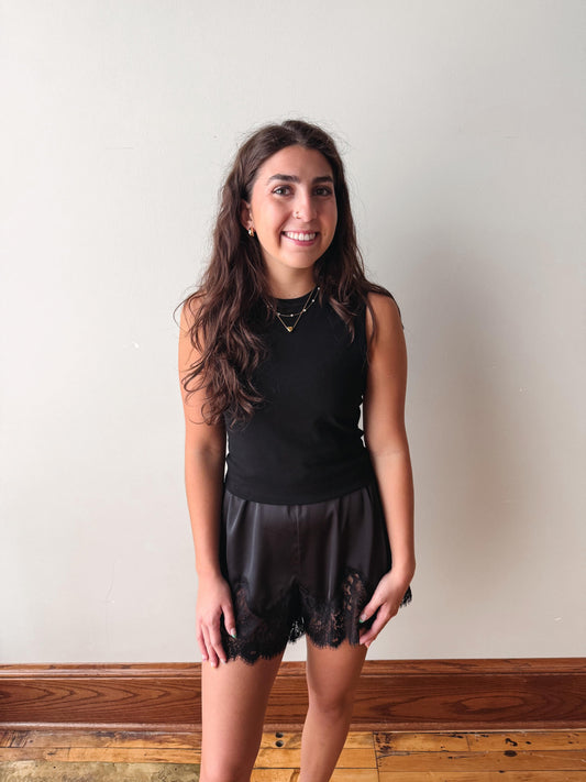 A young woman with long, wavy brown hair stands against a plain off-white wall wearing a black sleeveless top and black sheer lace shorts.