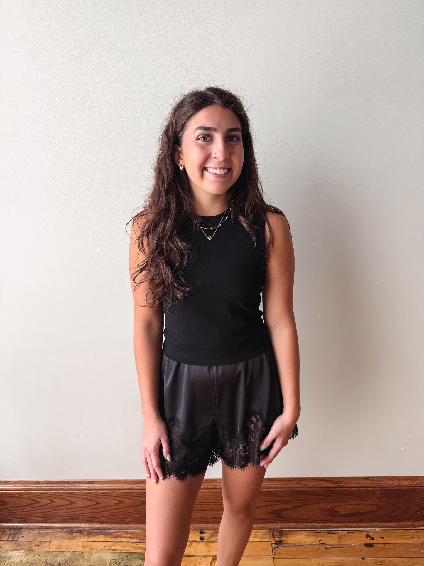A young woman with long, wavy brown hair stands against a plain off-white wall wearing a black sleeveless top and black sheer lace shorts.