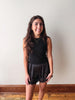 A young woman with long, wavy brown hair stands against a plain off-white wall wearing a black sleeveless top and black sheer lace shorts.