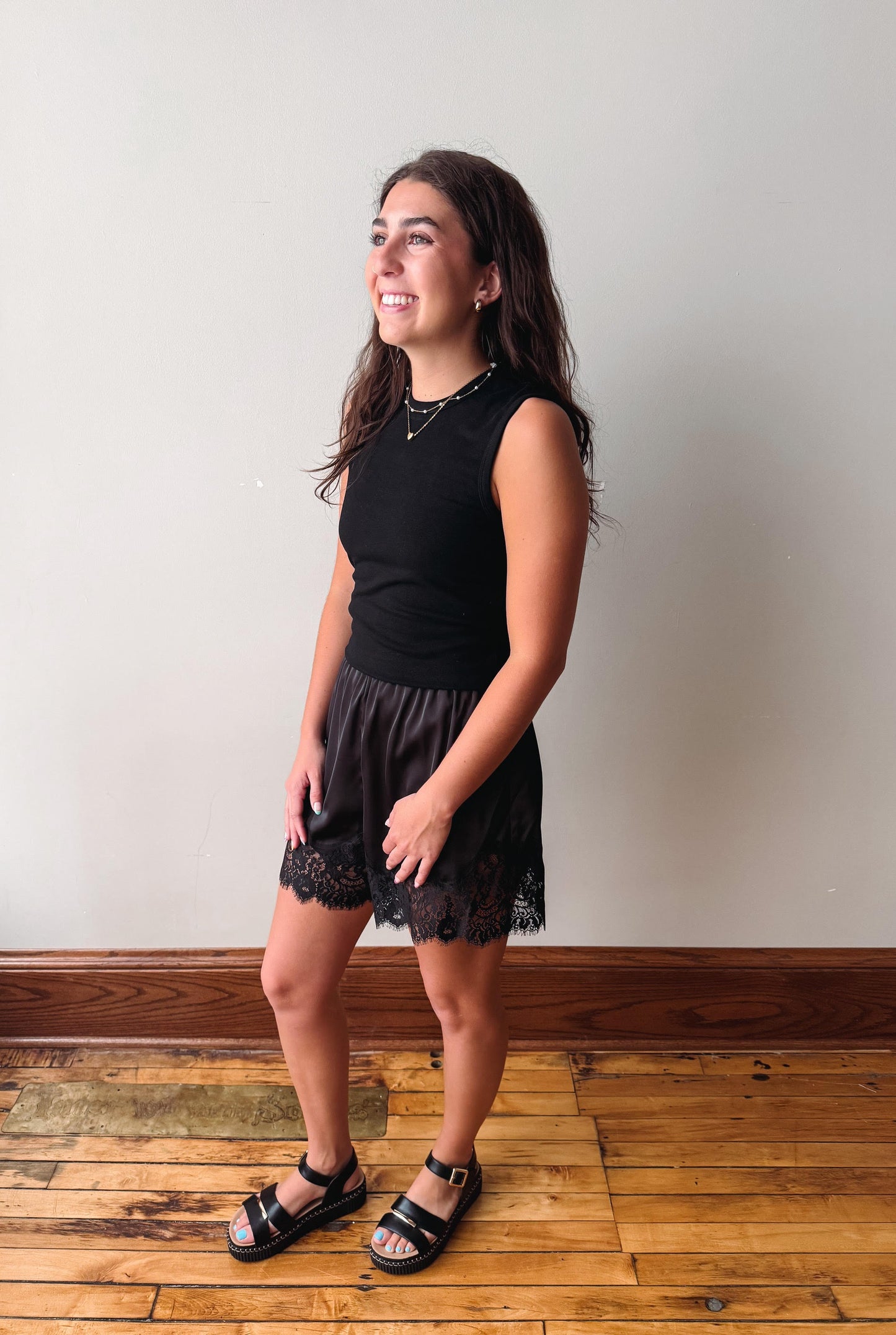 A young woman modeling a black sleeveless top and a black lace-trimmed skirt, paired with black slide sandals, standing against a plain light gray wall on a wooden floor.