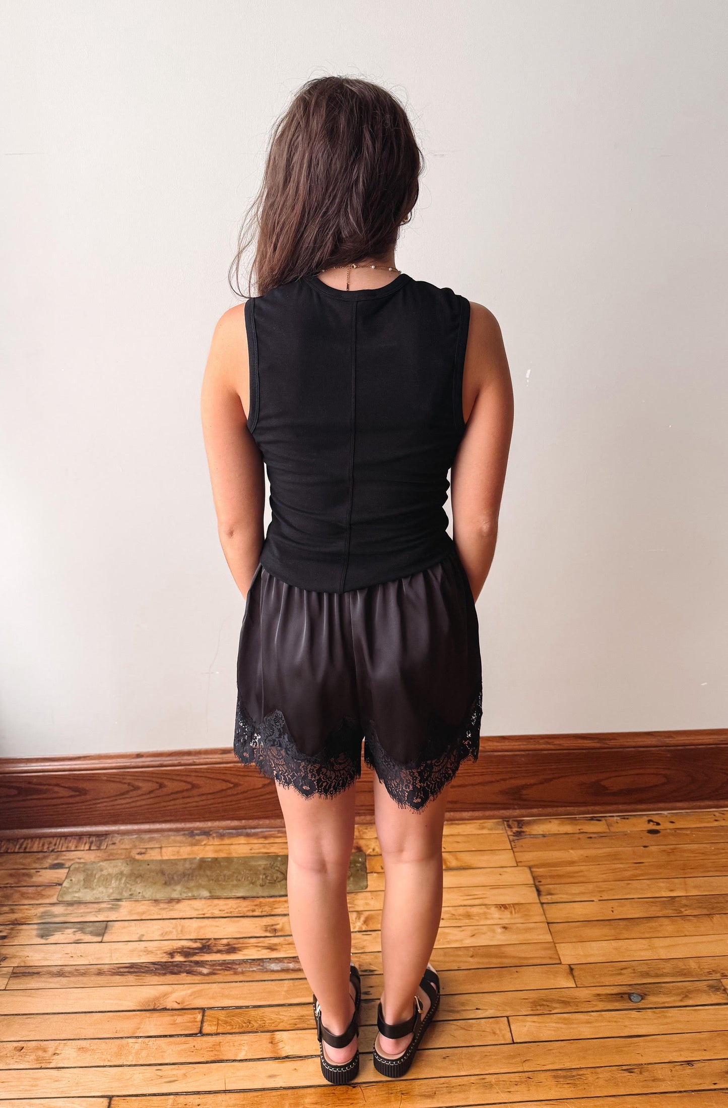A woman viewed from behind wearing a black sleeveless top and matching black lace-trimmed shorts, standing on a wooden floor against a plain light wall.