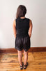 A woman viewed from behind wearing a black sleeveless top and matching black lace-trimmed shorts, standing on a wooden floor against a plain light wall.