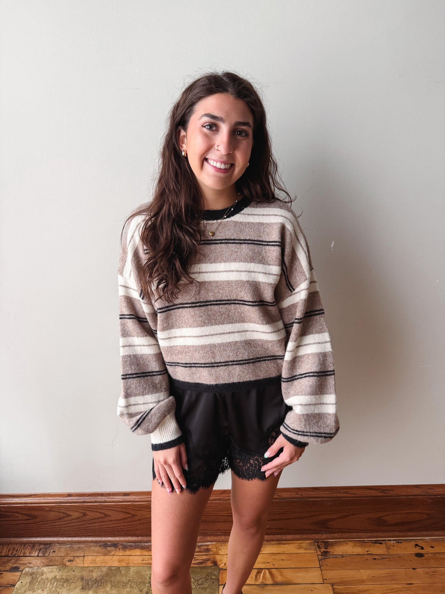 A woman modeling a cozy, long-sleeve striped knit sweater in beige, brown, and black, paired with black lace-up shorts, standing against a plain off-white wall with a wooden floor visible.