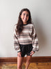 A woman modeling a cozy, long-sleeve striped knit sweater in beige, brown, and black, paired with black lace-up shorts, standing against a plain off-white wall with a wooden floor visible.