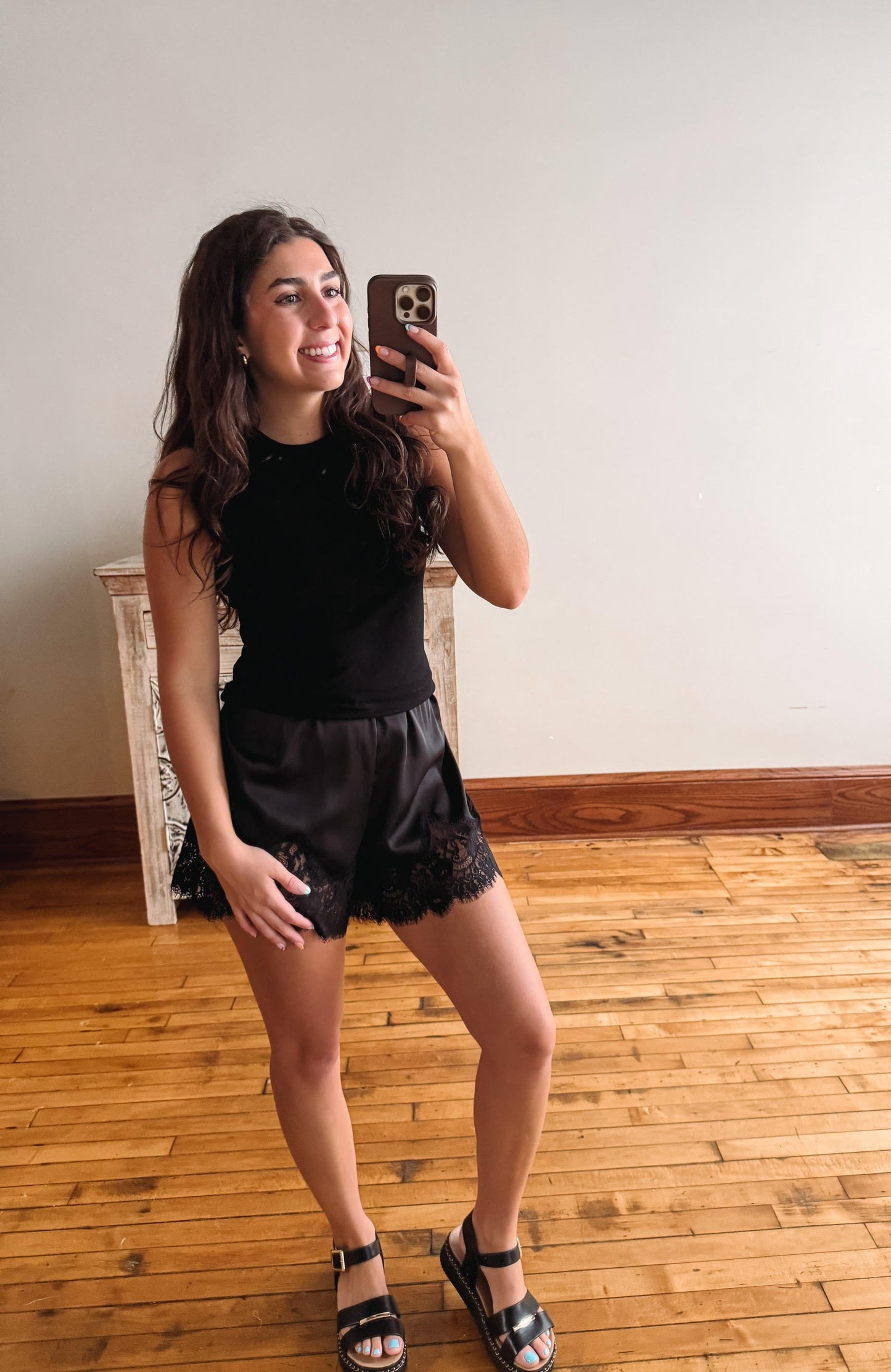 A woman takes a mirror selfie in an indoor room with light wood floors and a plain wall, wearing a black sleeveless top and black lace-trimmed shorts.