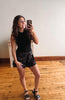 A woman takes a mirror selfie in an indoor room with light wood floors and a plain wall, wearing a black sleeveless top and black lace-trimmed shorts.