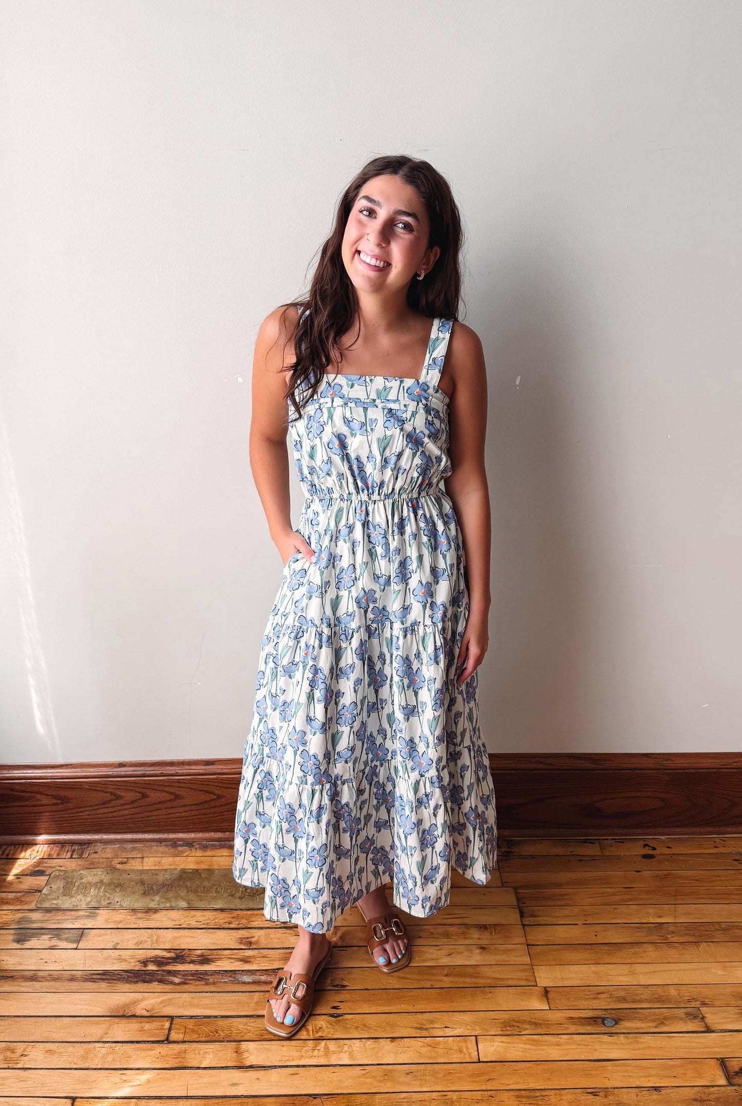 A woman modeling a sleeveless midi dress with a blue and white floral pattern, standing on a wooden floor against a plain off-white wall.