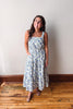 A woman modeling a sleeveless midi dress with a blue and white floral pattern, standing on a wooden floor against a plain off-white wall.