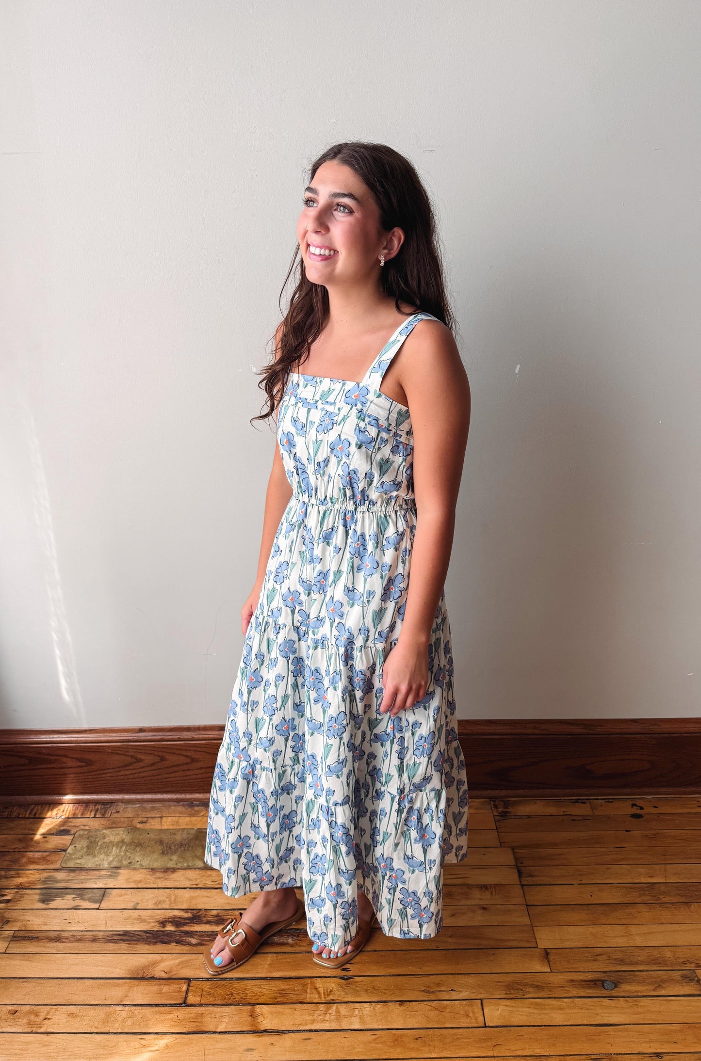 A woman wearing a blue and white floral print midi dress with a smocked bodice and thin shoulder straps, standing on a wooden floor against a plain wall.