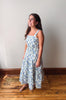 A woman wearing a blue and white floral print midi dress with a smocked bodice and thin shoulder straps, standing on a wooden floor against a plain wall.