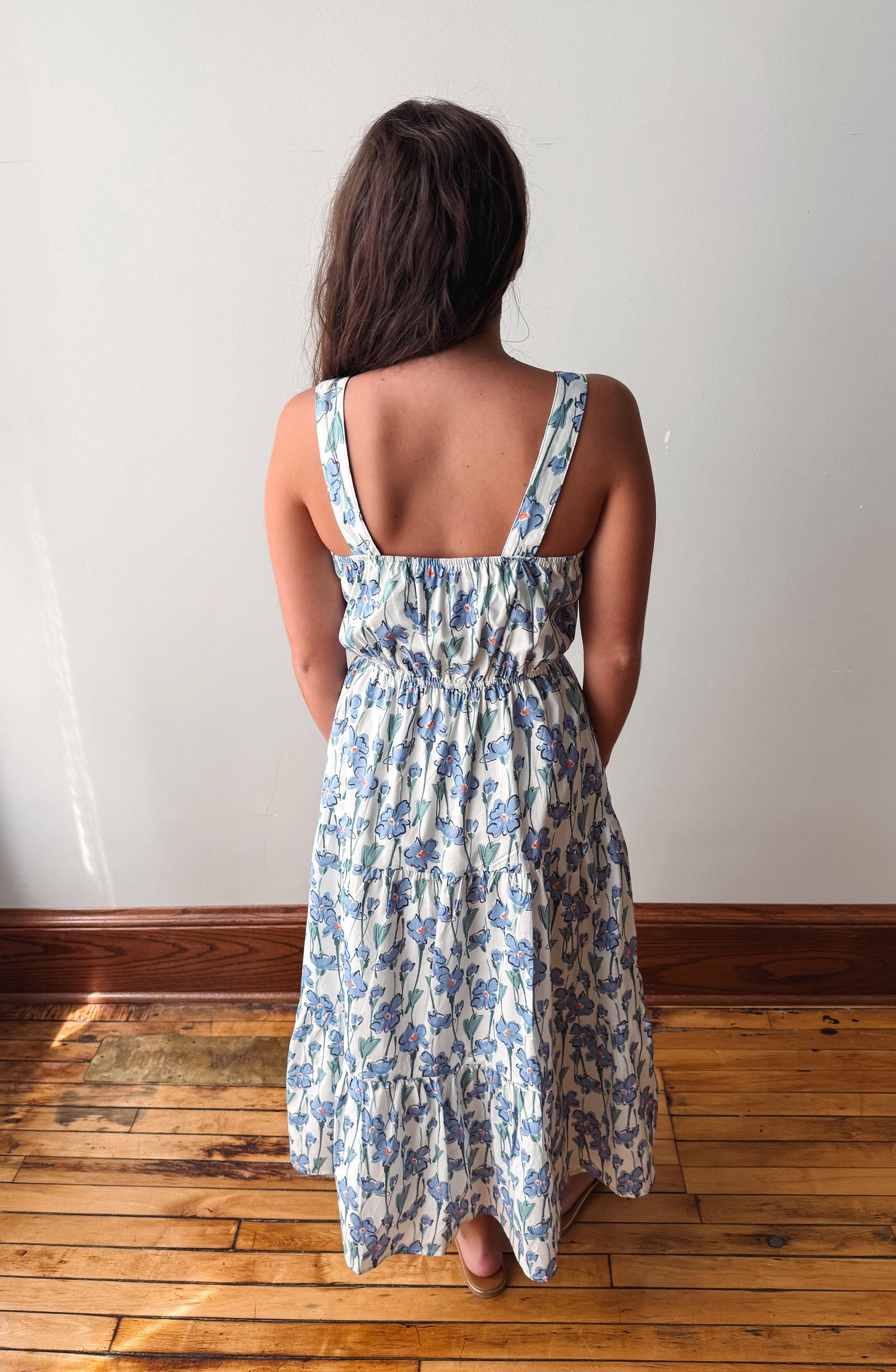 A woman viewed from behind wearing a sleeveless, tiered midi dress with a blue floral pattern on a white background, standing on a wooden floor against a plain white wall.